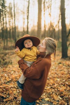 a woman holding a baby in her arms while standing in the woods with leaves on the ground