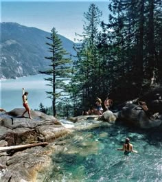 several people are swimming in the water near some rocks and pine trees with mountains in the background