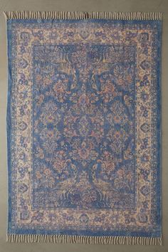 an antique rug with fringes on the bottom and blue ground, in front of a gray wall