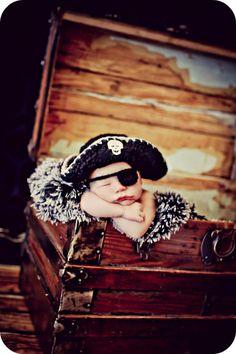 a baby in a pirate hat sitting on top of an old wooden trunk with his hands under his chin