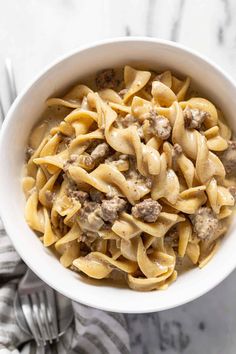 a white bowl filled with pasta and meat on top of a marble table next to silverware