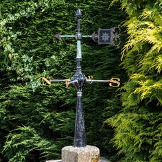 a weather vane on top of a stone pillar in front of some green bushes and trees