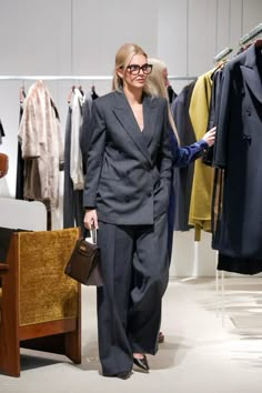 a woman in a suit is walking through a clothing store while holding a purse and looking at the camera