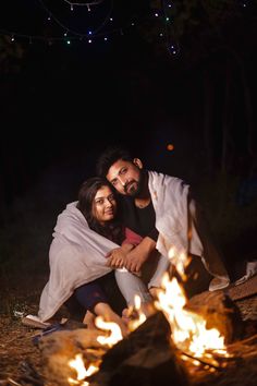 a man and woman sitting in front of a campfire with blankets over their heads