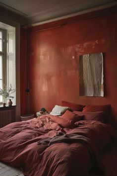 an unmade bed in front of a window with red walls and wood floors,