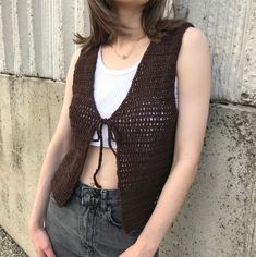 a woman standing next to a wall wearing a brown crocheted vest and jeans