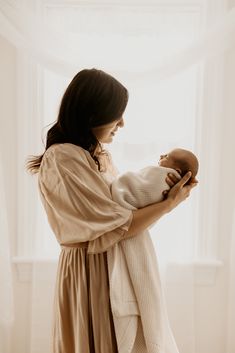 a woman holding a baby in her arms and looking out the window with sunlight streaming through