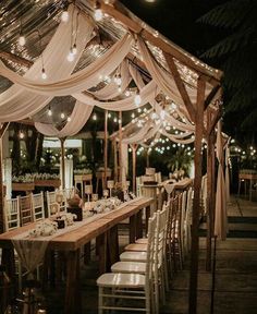 a long table with white chairs under a canopy covered in lights and draping