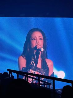 a woman singing into a microphone in front of a large screen with an image of herself on it