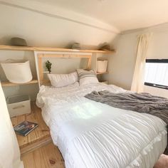 a white bed sitting under a window next to a wooden shelf filled with pillows and blankets