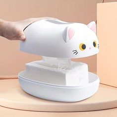 a white cat shaped tissue dispenser sitting on top of a table next to a person's hand