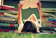 a woman laying on the grass reading a book with books stacked up in front of her