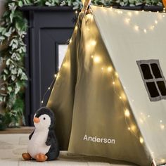 a penguin sitting in front of a teepee tent with lights on it and a stuffed penguin next to it