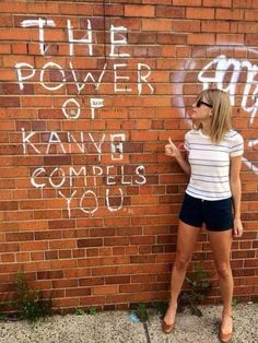 a woman standing in front of a brick wall with graffiti on it and pointing to the side