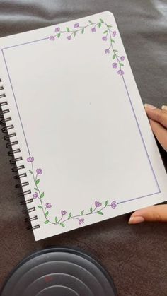 a hand holding a spiral notebook with flowers on the cover and blank paper in front of it