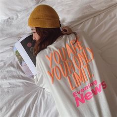 a woman laying on top of a bed with a book in her hand and wearing a beanie