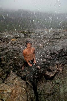 a shirtless man standing on top of a rocky cliff covered in water and rain