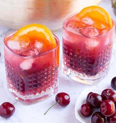 two glasses filled with cherries and oranges on a marble table next to a bowl of cherries