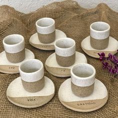 six white cups and saucers sitting on top of a cloth covered tablecloth next to a purple flower