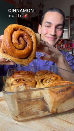 a man holding up a cinnamon roll in front of a glass dish on a wooden table