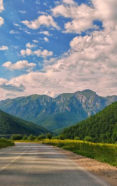 an empty road in the middle of mountains under a blue sky with fluffy white clouds