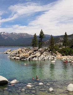 people are swimming in the water near some rocks and trees with mountains in the background