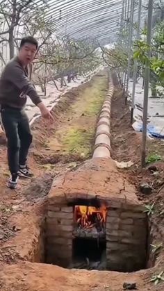 a man standing next to a brick oven in the middle of a dirt field with trees