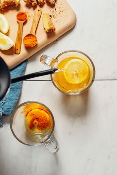 two glasses filled with liquid next to sliced lemons and spices on a cutting board