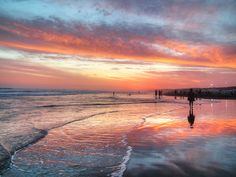 people walking on the beach at sunset with surfboards in the water and colorful clouds