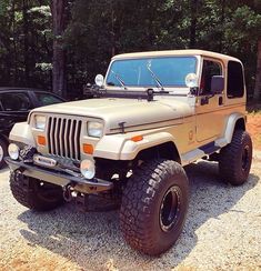 a tan jeep parked next to a black car in front of some trees and bushes