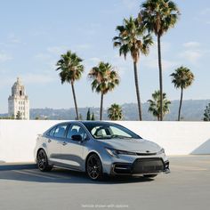 a silver car is parked in front of some palm trees and a white wall with a clock tower behind it