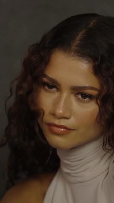 a woman with curly hair wearing a white turtle neck top and looking at the camera