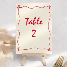 a table number sign sitting on top of a wooden easel next to gold forks