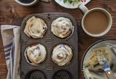 a muffin tin filled with cupcakes on top of a wooden table next to two cups of coffee