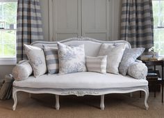 a white couch with many pillows on it in front of two windows and a table