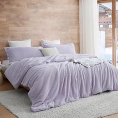 a bed with purple comforter and pillows on top of it in front of a wooden wall