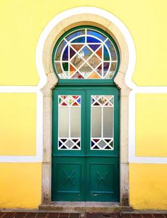 a green door with stained glass in front of a yellow and white building on a brick sidewalk