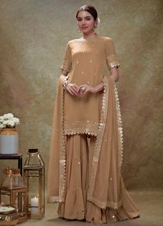 This exquisite Beige Georgette Kurta Sharara Set is a perfect blend of elegance and tradition. Made from georgette fabric, the beige kurta is adorned with delicate embroidery featuring mirrors, gota, sequins, and pearls, creating a stunning visual appeal. The neckline, hem, and sleeves are intricately embellished, complemented by small floral buttis that add a touch of grace. Teamed with a tiered sharara that showcases scattered mirror work, this ensemble exudes sophistication. The look is completed with a matching net dupatta, beautifully bordered and dotted with buttis, making it an ideal choice for Home Pooja, family gathering or festive occasions. Composition : Kurta and Sharara - Viscose Georgette and Dupatta - Soft net Care: Dry Clean Only and Vacuum Storage This product can be custo Latest Sharara Designs Party Wear, Golden Sharara, Beige Sharara, Mirror Work Kurta, Bridal Crop Top, Beige Kurta, Kaftan Kurta, Georgette Kurta, Kurta Lehenga