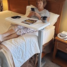 an older woman laying in bed while looking at her cell phone and holding a cup