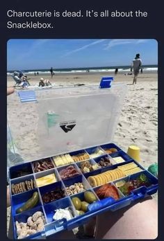 someone is sitting on the beach with their lunch box in front of them and there are people walking around