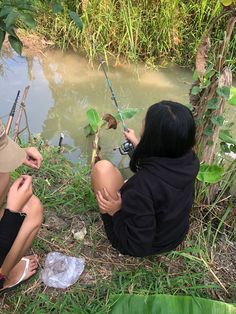 two people sitting on the ground next to a body of water with fishing rods in their hands