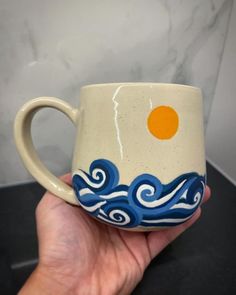 a hand holding a coffee cup with an orange on top and blue waves in the bottom