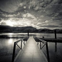 a long wooden dock sitting on top of a lake under a cloudy sky with mountains in the background