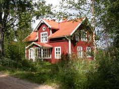 a red house is surrounded by trees and grass on the side of a dirt road