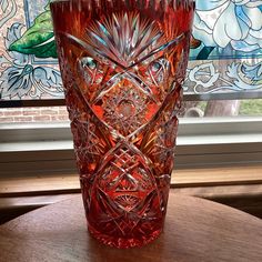 a red glass vase sitting on top of a wooden table next to a stained glass window