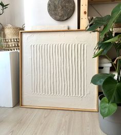 a white framed art piece sitting on top of a wooden floor next to a potted plant