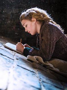 a woman sitting on the floor writing with a pen in her hand and looking at an open book