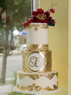 a three tiered white and gold wedding cake with red flowers on the top layer