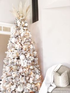 a white christmas tree decorated with silver and gold ornaments in a living room next to a couch