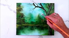 a person holding a pencil in their hand near a painting on the wall with water and trees